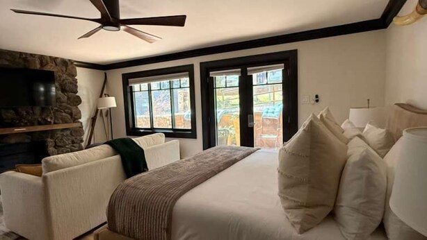 Une chambre à coucher avec un lit, un ventilateur de plafond, un mur en pierre et une vue dégagée sur l’extérieur grâce à de grandes fenêtres.