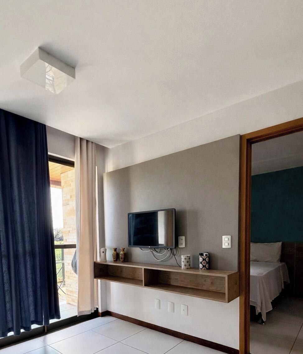 Une chambre d’hôtel avec un téléviseur fixé au mur, une étagère en bois en dessous et un lit recouvert d’une couverture blanche.