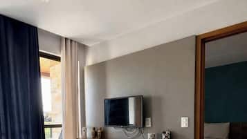 Quarto de hotel com televisão montada na parede, prateleira de madeira abaixo e cama com colcha branca.