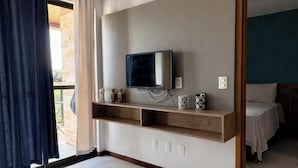 A hotel room with a television on a wooden shelf, a bed, and a view of the outdoors. - Flat 106 S Iloa Residence - Barra de São Miguel, Alagoas, vacation rental (Barra de São Miguel)