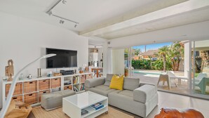 Ein modernes Wohnzimmer mit einem großen Flachbildfernseher, einem grauen Sofa mit gelbem Kissen, Weidenkorbmöbeln und einer Glasdoor zu einem Poolbereich.
