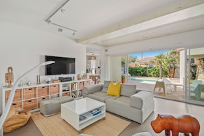 Una sala de estar moderna con un televisor de pantalla plana grande, un sofá gris con un cojín amarillo, almacenamiento de mimbre y un ventanal que da a una zona de piscina.
