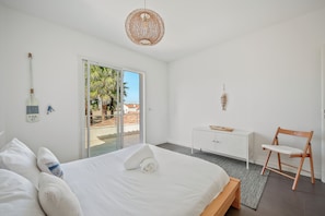 Habitación de hotel moderna con cama, escritorio, silla, lámpara colgante y vista a palmeras a través de la puerta.