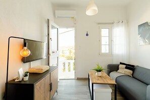 A modern living room with a sofa, a TV mounted on a cabinet, a wooden table with a plant, and a ceiling light. - Harbor View Apartment in Mykonos Town (Mykonos)