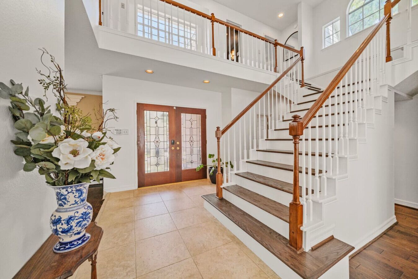 Un hall espacioso con una escalera de madera, un par de puertas dobles con paneles de vidrio y un jarrón con flores sobre una mesa de madera.