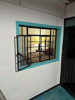 Ein Fenster mit Metallgitter, ein Bett mit gestreifter Bettdecke und eine Lampe im Hintergrund.