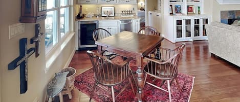 Un comedor con una mesa y sillas de madera, una cocina al fondo y una estantería llena de objetos varios.