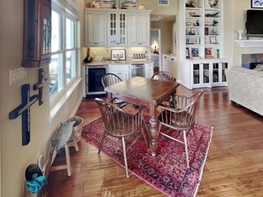 A dining area with a wooden table and chairs, a kitchen in the background, and a bookshelf filled with various items. - Galveston Baywatch (Galveston)