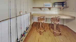 A room with a wooden floor, two wicker chairs, a white wall with fishing rods, and a shelf with a white air conditioner. - Galveston Baywatch (Galveston)