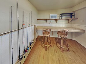 Une pièce avec un plancher en bois, deux fauteuils en osier, un mur blanc orné de cannes à pêche et une étagère sur laquelle se trouve un climatiseur blanc.