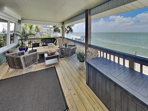 A covered outdoor deck with a wooden table and chairs, a small rug, and a view of the ocean. - Galveston Baywatch (Galveston)