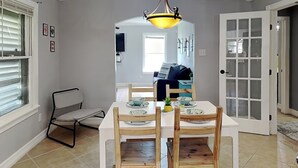 A dining area with a white table, wooden chairs, a yellow pendant light, and a view into the living room through a glass door. - Shell City (Galveston)