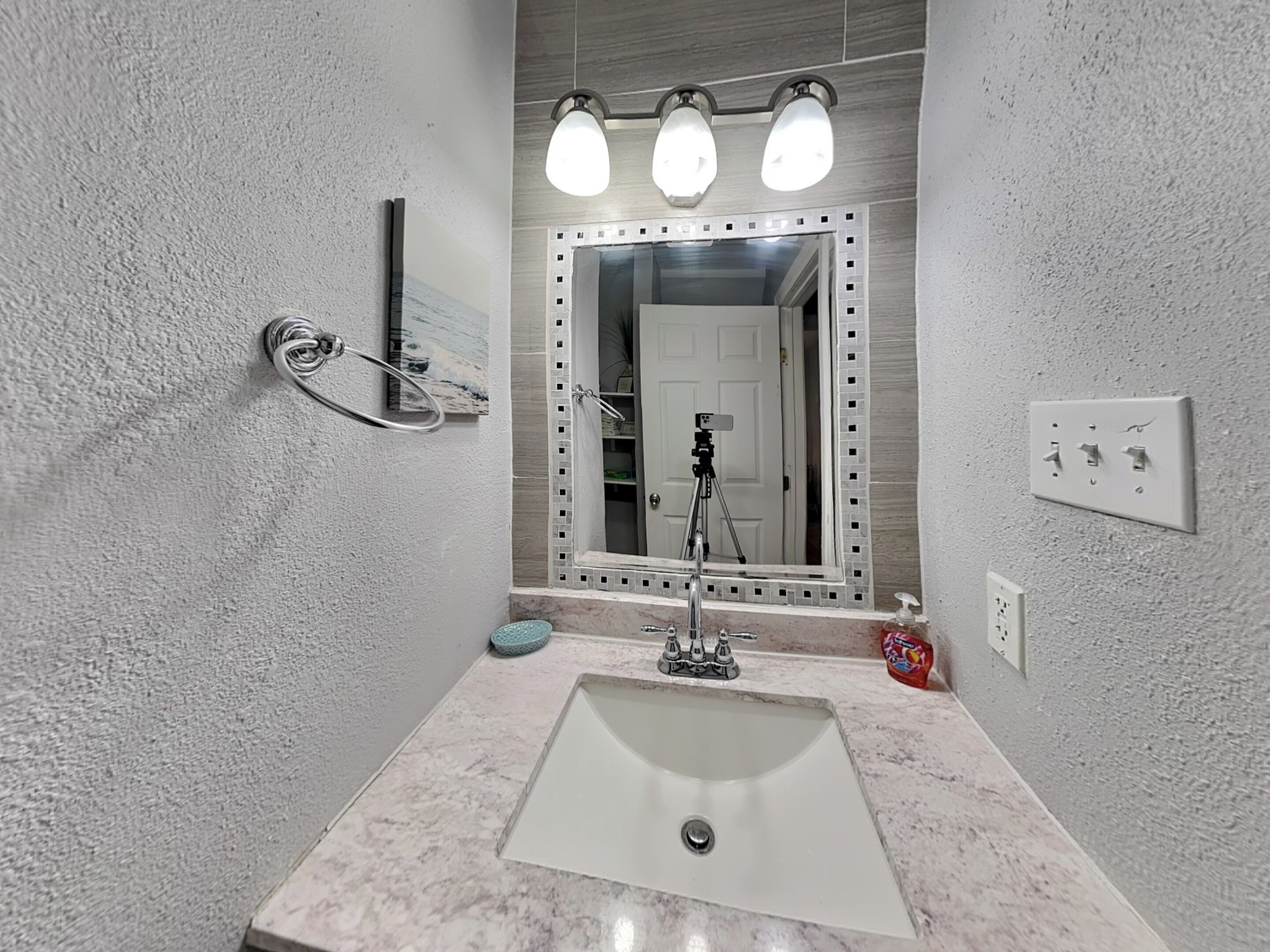 A bathroom with a marble sink, a large mirror with lights, a towel rack, and a tripod.