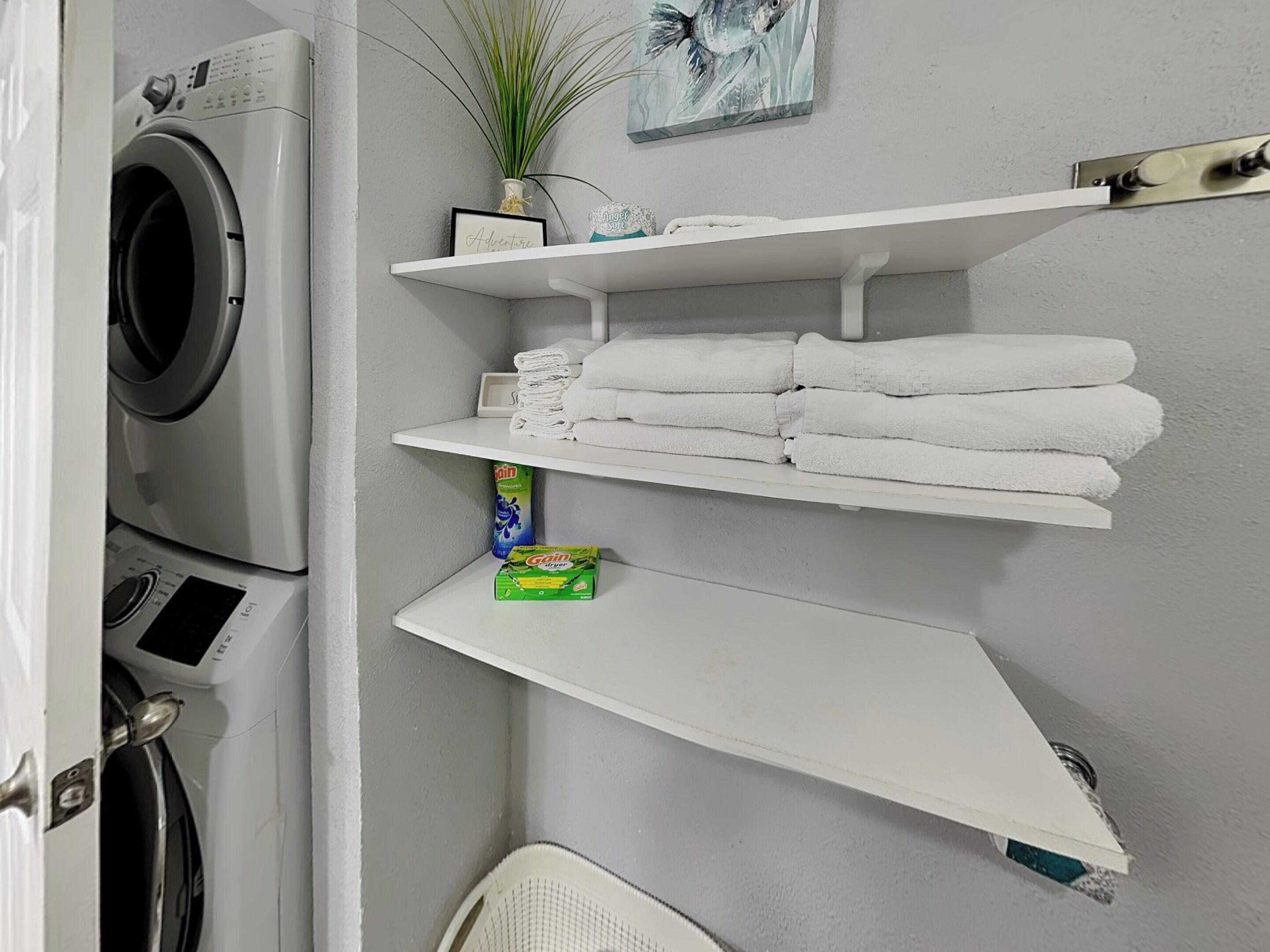 Een wasruimte met een wasmachine, planken met opgevouwen handdoeken, een plant en een lijst met een foto.