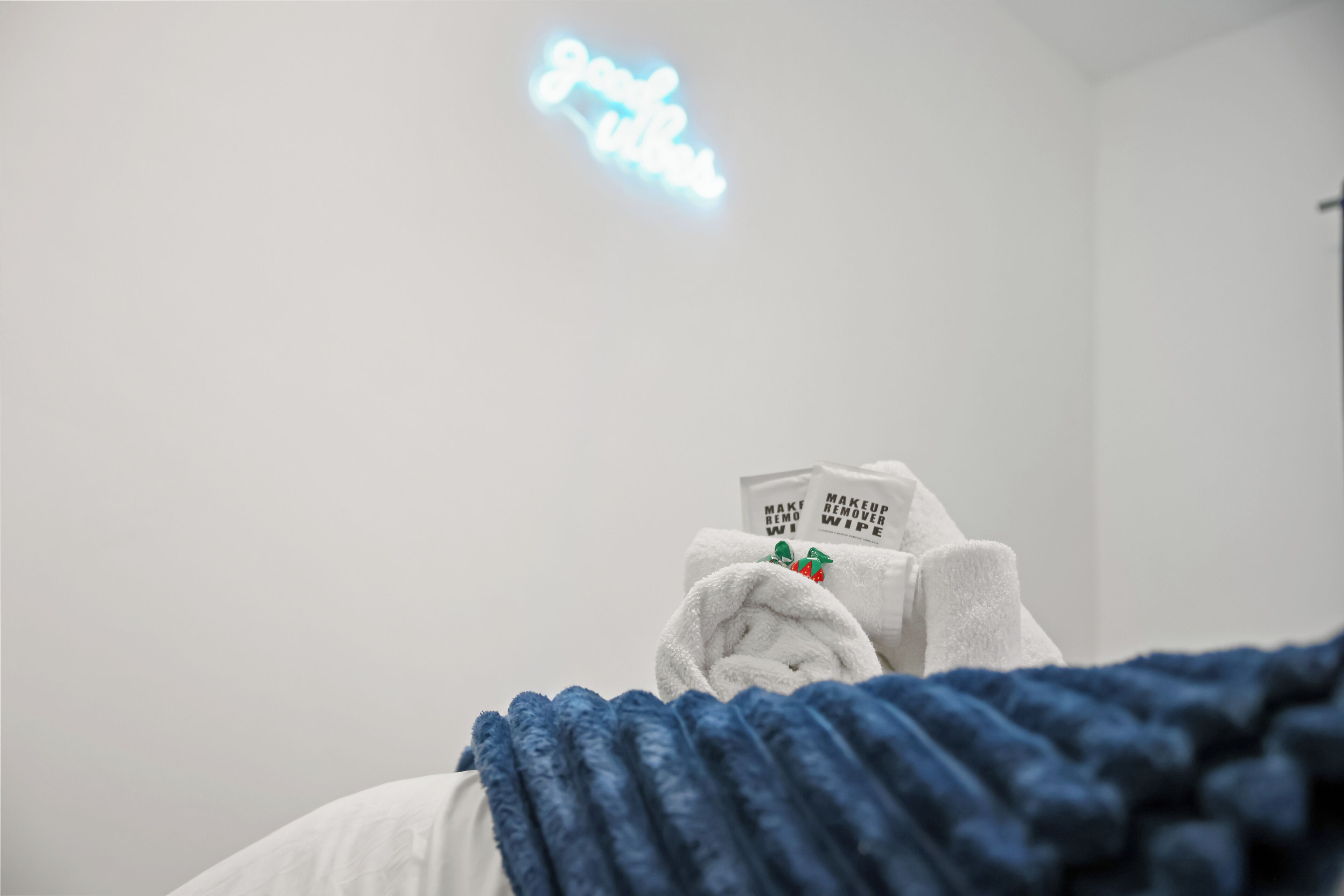 Ett hotellrum med en säng täckt av ett blått täcke, en vit kudde och en vit handduk med en grön logotyp. På väggen finns en neonskylt med texten ”Bli frisk”.