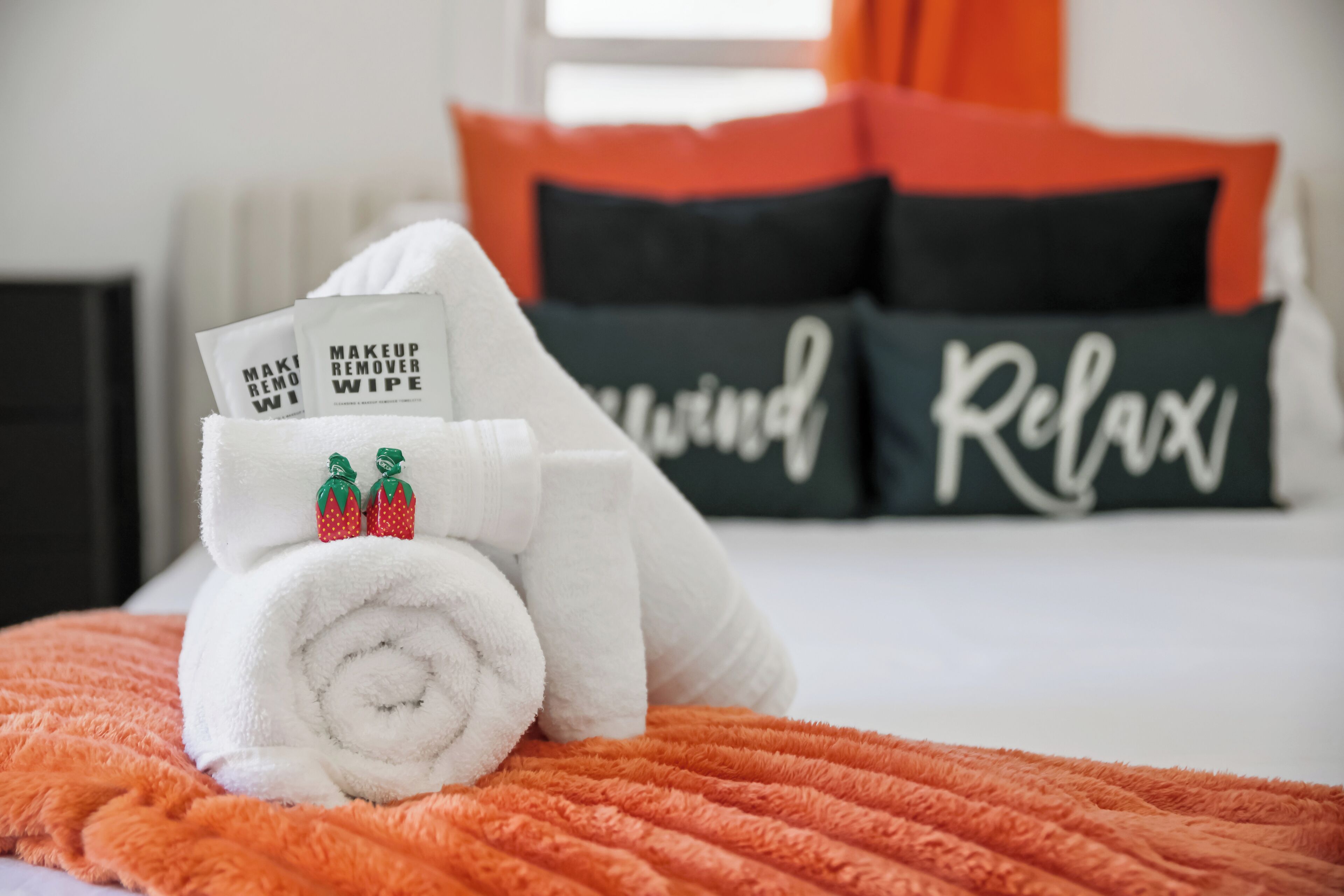 A neatly made bed with a rolled white towel and two makeup remover wipes, a black pillow with 'Relax' written on it, and a black pillow with 'Relax' written on it.