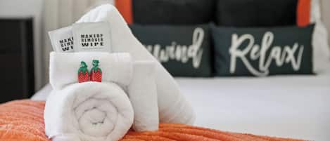 A neatly made bed with a rolled white towel and two makeup remover wipes, a black pillow with 'Relax' written on it, and a black pillow with 'Relax' written on it.