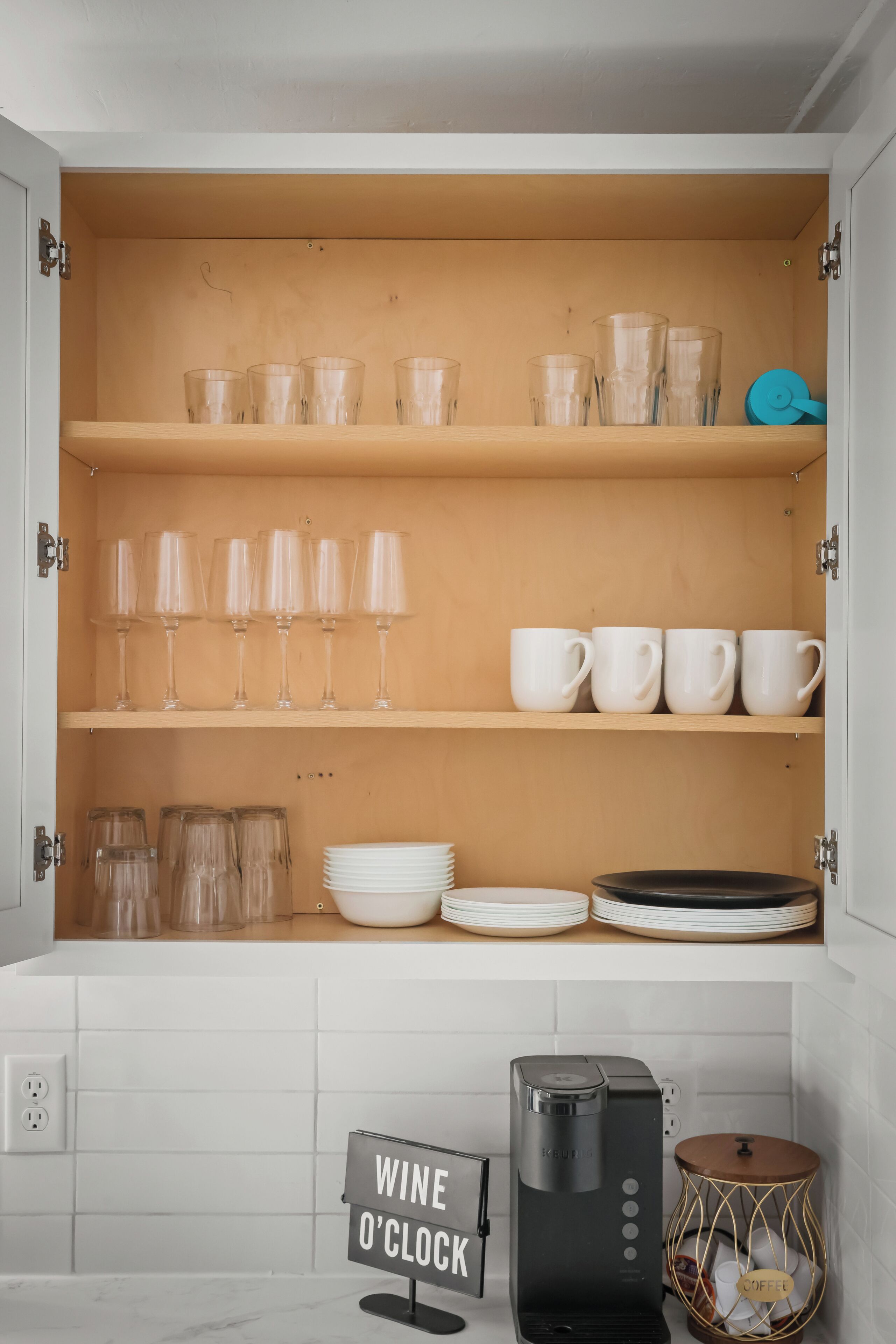 A kitchen with a wine and coffee station, clear glassware, and white mugs on shelves.