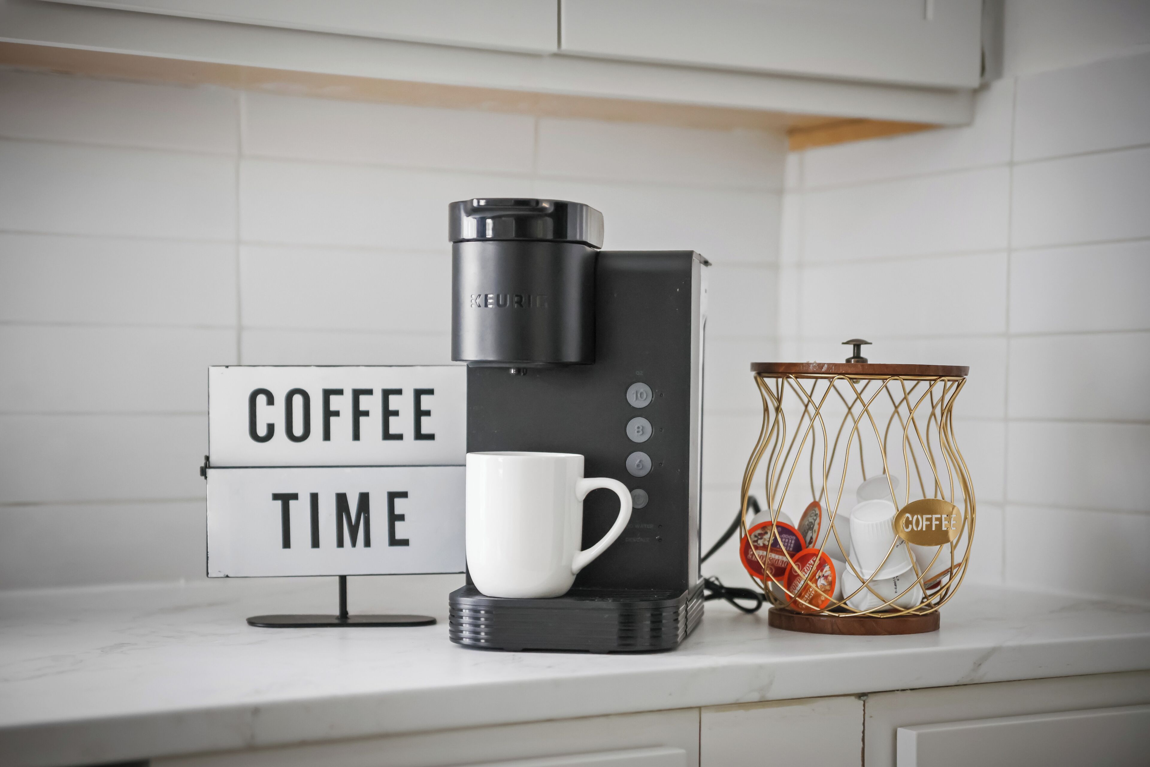 Une machine à café, une tasse blanche et un panneau indiquant « PAUSE CAFÉ » sur un plan de travail de cuisine.