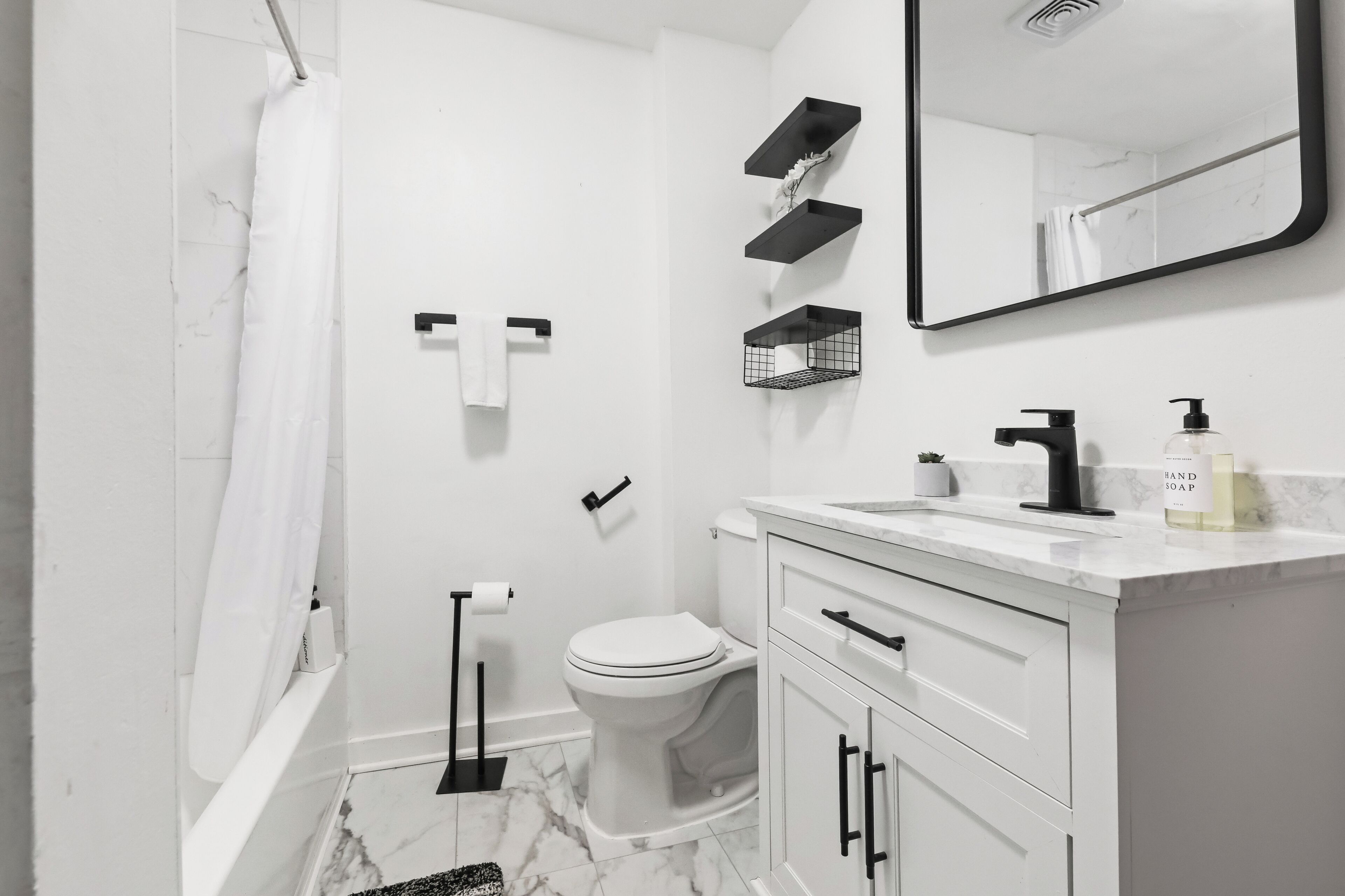 Un baño moderno con inodoro blanco, cortina de ducha, toallero, dispensador de papel higiénico, lavamanos con espejo y dispensador de jabón.