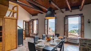A dining area with a table set for four, a fruit bowl, wine, and a view of mountains through large windows. - Casa Tranquillità Lario, Plesio, Italy (Plesio)