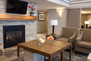 A hotel room with a fireplace, a flat-screen TV mounted above it, a sofa, a table with flowers, and a lamp. - 1BR King Suite w/ Kitchen | Free Breakfast + Gym (Vaughan)