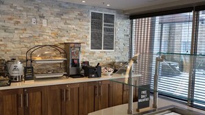 A breakfast bar with a stone wall backdrop, a coffee machine, and a glass display case with food items. - 1BR King Suite w/ Kitchen | Free Breakfast + Gym (Vaughan)