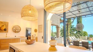 Una cocina moderna con una isla grande, lámparas colgantes de mimbre y vistas a una piscina y palmeras.