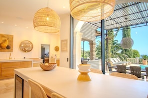 A modern kitchen with a large island, wicker pendant lights, and a view of a pool and palm trees. - 🌅 Cavalaire | Infinite Sea View & Infinity Pool (Cavalaire-sur-Mer)
