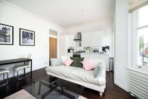 Ein modernes Wohnzimmer mit einem Glastisch, einer Couch mit Kissen, einem Essbereich mit einem schwarzen Tisch und Barhocker sowie einer Küche mit weißen Schränken und einem Herd.