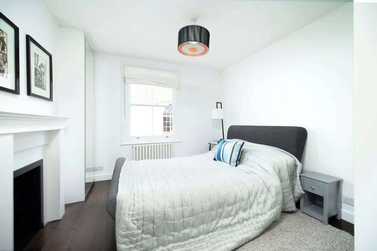 A bedroom with a bed, a nightstand, a lamp, a fireplace, and a window.