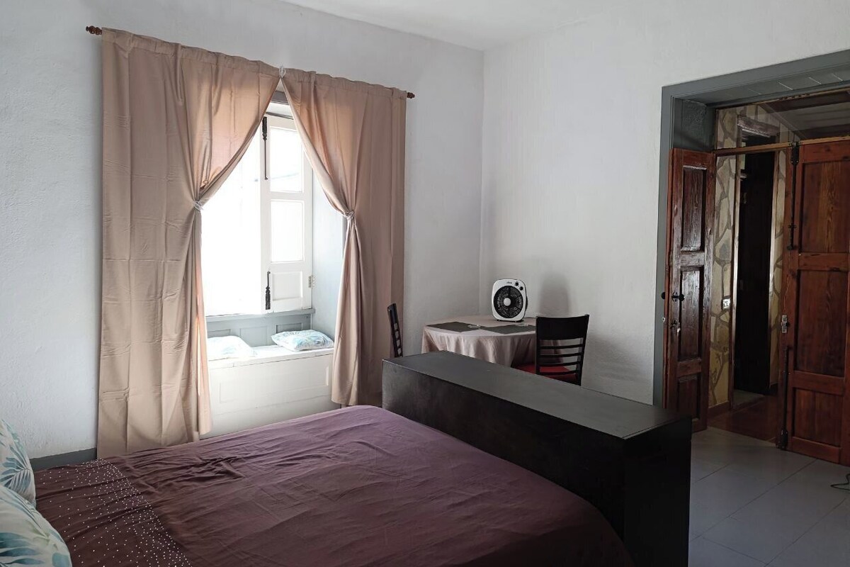 A bedroom with a bed, a desk with a fan, and a window with curtains.