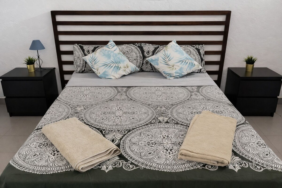 A bed with a patterned bedspread, two pillows, two bedside tables with lamps, and a small plant on each table.