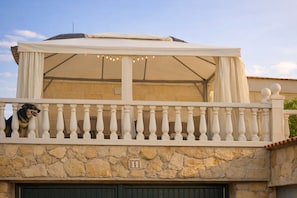 Un bĂątiment en pierre dotĂ© dâun balcon, dâun espace extĂ©rieur couvert amĂ©nagĂ© en coin salon et dâun chien sur la rambarde.