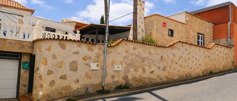 A street view with a stone wall, a white building with a balcony, and a red building with a flat roof.