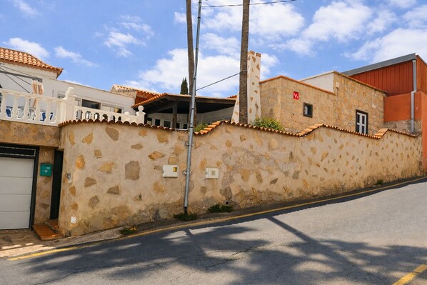A street view with a stone wall, a white building with a balcony, and a red building with a flat roof. - Zakshouse (Arona)