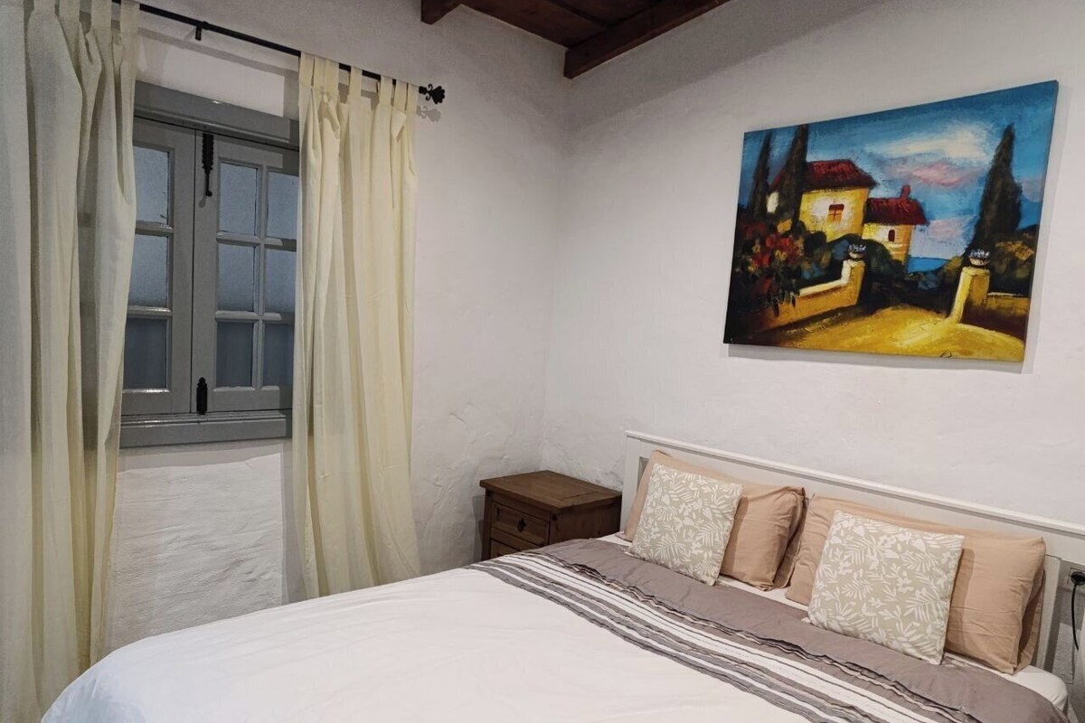 A bedroom with a bed, a nightstand, a window with curtains, and a painting on the wall.