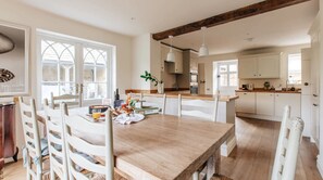 A dining area with a wooden table, white chairs, and a kitchen in the background. - 4-Bed Cotswolds Getaway     Perfect for Families (Kingham)
