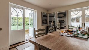 A dining area with a wooden table, chairs, and a view of a garden through open doors. - 4-Bed Cotswolds Getaway     Perfect for Families (Kingham)
