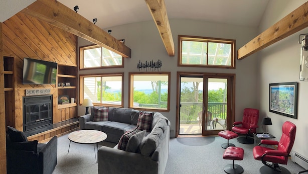 A cozy living room with a fireplace, a grey sofa, red chairs, and large windows offering a view of the outdoors. - Okemo Trailside: 3BR + Bonus Room, Hot Tub, Sauna & On-Demand Shuttle to Sachem (Ludlow)