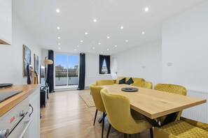 Un coin repas avec une table en bois et des chaises jaunes, une cuisine équipée d’appareils modernes et une vue dégagée grâce à de grandes fenêtres.