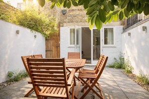 Une terrasse avec une table et des chaises en bois, un mur blanc et une porte donnant accès à un bâtiment.