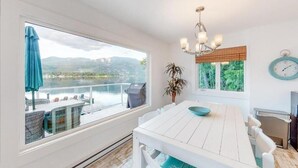 Een eethoek met een tafel en stoelen, een raam met uitzicht op een meer en bergen, een plant en een klok aan de muur.