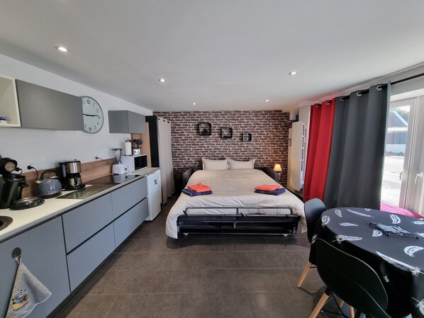 A compact hotel room with a kitchenette, a bed with pillows, a desk, and a chair.
