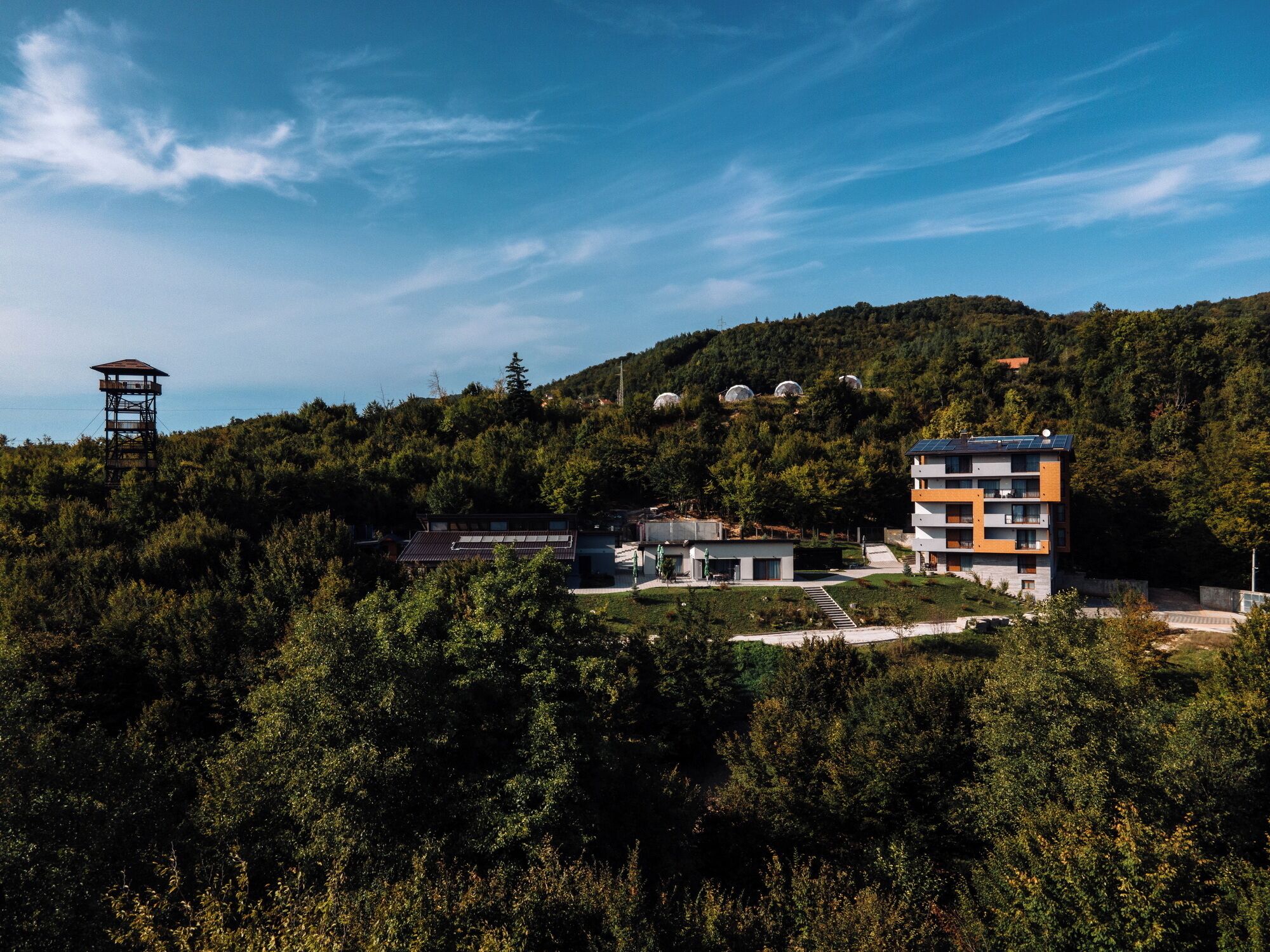 A modern building with a flat roof, surrounded by dense forest and hills.
