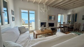 A spacious living room with a sofa, a coffee table, a fireplace, and a dining area with chairs and a table. - Panoramic Sea View Villa Near Beach and Town (Mykonos)