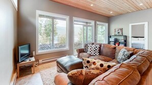 Una sala de estar con un sofá de cuero marrón, un televisor sobre un mueble bajo y amplios ventanales con vistas a la vegetación.
