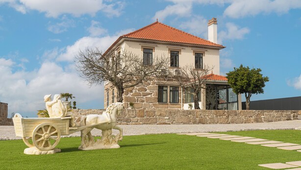 Un edificio de piedra con tejado de tejas rojas, una pared de piedra y un césped con un carruaje tirado por caballos.