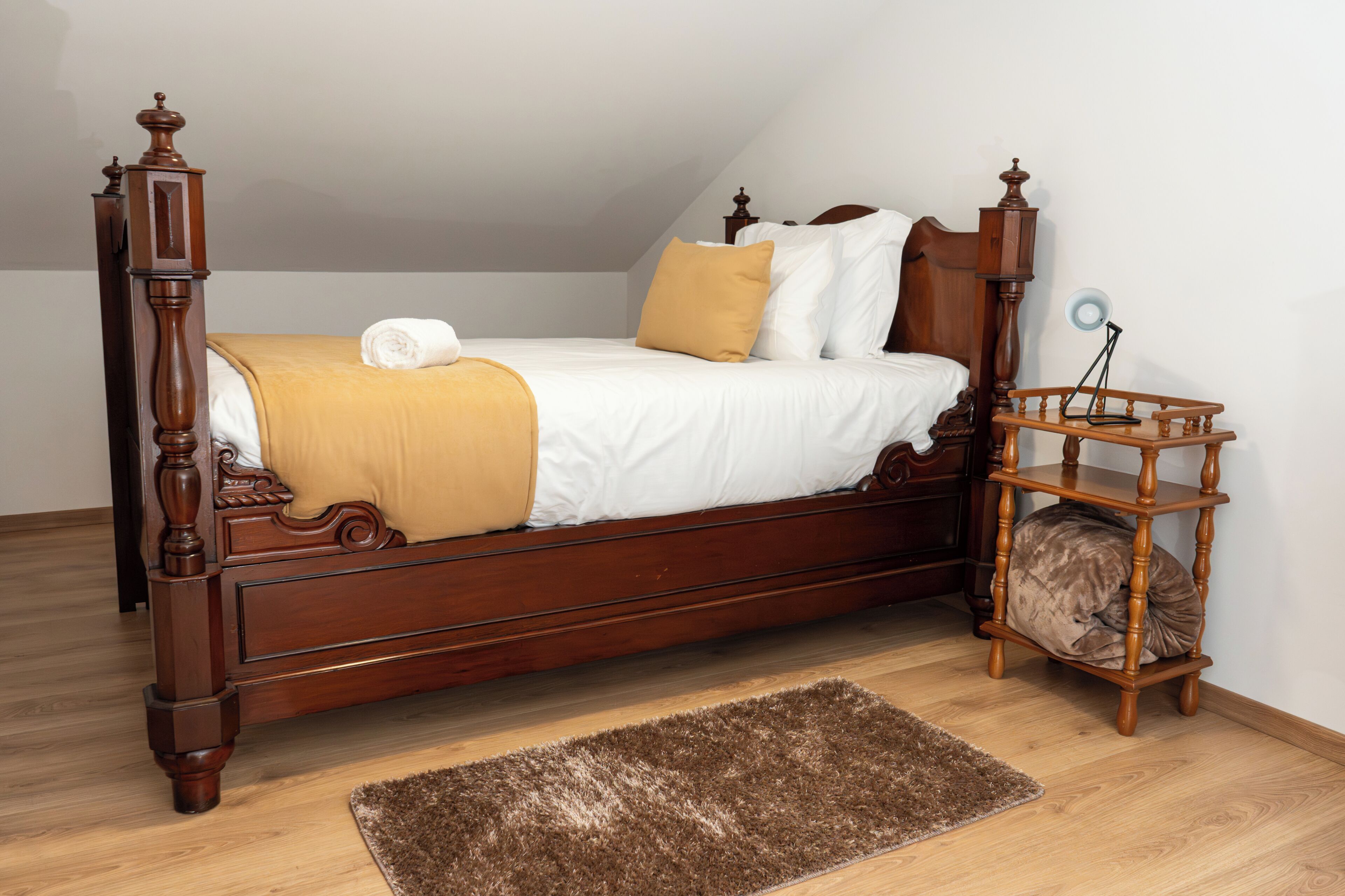 Una cama con dosel de madera, con una colcha blanca y amarilla, un tapete marrón y una mesita de noche de madera con una lámpara.
