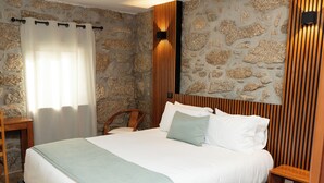 Una cama con cabecera, una mesita de noche de madera, una silla y un muro de piedra.